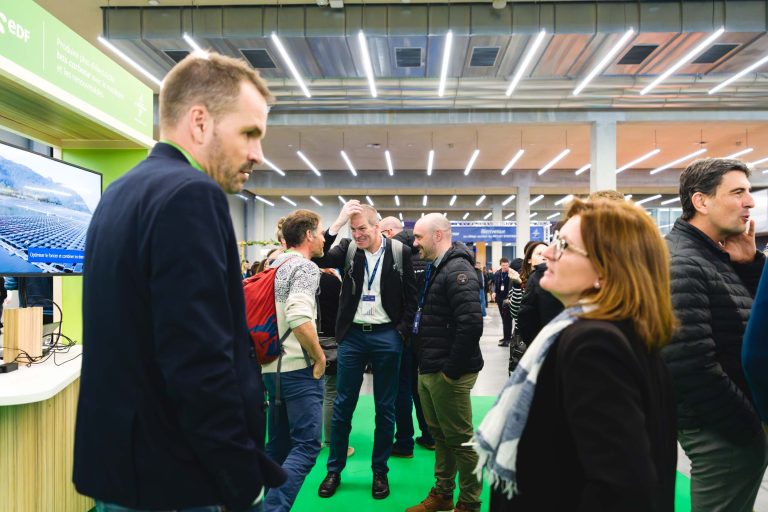 Un groupe de personnes en tenue décontractée converse et tisse des liens lors d'un événement ou d'une conférence en salle. Certains portent des cordons et des badges. L'espace est doté de hauts plafonds, de lumières vives et d'un sol vert. Des cabines et des écrans sont visibles à l'arrière-plan.