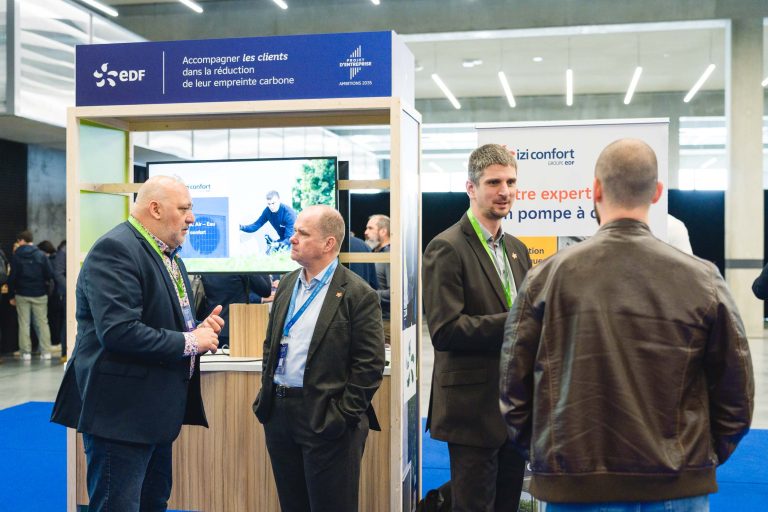 Quatre hommes en tenue de travail discutent près d'un stand EDF bleu et blanc lors d'un événement professionnel. Derrière eux se trouvent des panneaux d'information et un écran de télévision montrant un homme à vélo, avec en arrière-plan une salle moderne et spacieuse.