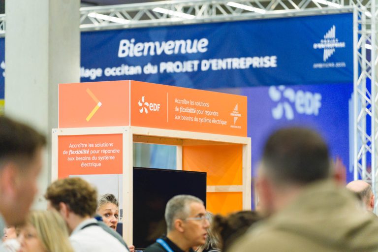 Une foule se tient près d'un stand EDF avec un panneau orange indiquant "Accroître les solutions de flexibilité pour répondre aux besoins du système électrique". Une bannière bleue portant les inscriptions "Bienvenue" et "PROJET D'ENTREPRISE" est suspendue à l'arrière-plan.