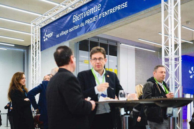 Un groupe de personnes en tenue d'affaires converse autour d'une table haute lors d'un événement en intérieur. Une bannière bleue indique "Bienvenue au village occitan du PROJET D'ENTREPRISE". Le cadre est moderne, avec des lumières et des murs en verre en arrière-plan.