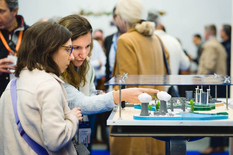 Deux femmes examinent attentivement une maquette détaillée d'une installation industrielle avec des cheminées et des tours de refroidissement et en discutent. Elles sont entourées d'autres personnes dans un environnement intérieur animé, suggérant un événement professionnel ou éducatif.