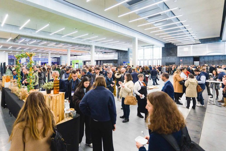 Une foule de personnes se mêle à une salle spacieuse, moderne et bien éclairée. Des tables avec de la nourriture et des décorations sont disposées sur la gauche, et les participants discutent, mangent et se promènent en tenue de ville.