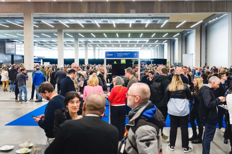 Un grand groupe de personnes est rassemblé dans une salle spacieuse et moderne, avec de hauts plafonds et un éclairage lumineux, engageant des conversations et tenant des boissons et des assiettes. Plusieurs personnes se tiennent debout sur un tapis bleu et des panneaux d'information sont visibles à l'arrière-plan.