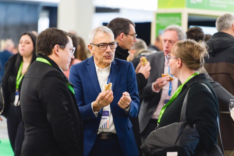 Un groupe de personnes habillées professionnellement et portant des cordons verts se réunit à l'intérieur, discutant et mangeant des amuse-gueules lors de ce qui semble être une conférence ou un événement de réseautage. Un homme en costume bleu fait des gestes animés tout en parlant avec deux autres participants.