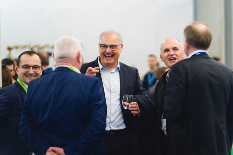 Un groupe de cinq hommes en costume se tient dans une pièce bien éclairée, souriant et riant ensemble. Un homme au centre tient un verre d'eau et fait des gestes en parlant. Des personnes et des décorations floues sont visibles à l'arrière-plan, créant une atmosphère animée.