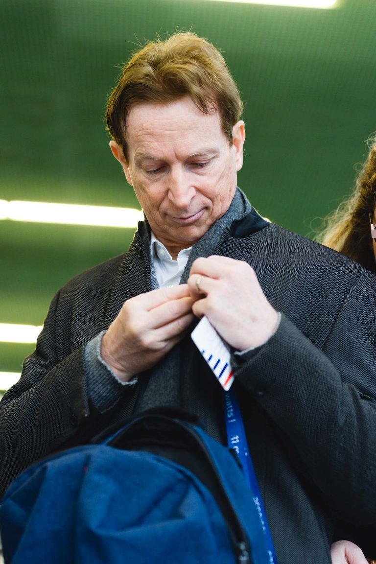 Un homme aux cheveux châtain clair, vêtu d'un manteau sombre sur une chemise blanche, regarde vers le bas tout en fermant son manteau. Il tient un cordon et des papiers. L'arrière-plan est verdâtre avec des lumières horizontales floues. Une autre personne est partiellement visible sur la droite.