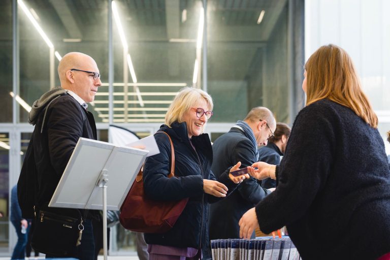 Une femme souriante, vêtue d'un manteau sombre, reçoit un badge d'un membre du personnel à un bureau d'enregistrement. Un homme à lunettes se tient à côté d'elle. D'autres personnes font la queue. Le cadre semble être un bâtiment moderne, très éclairé, avec des murs en verre.