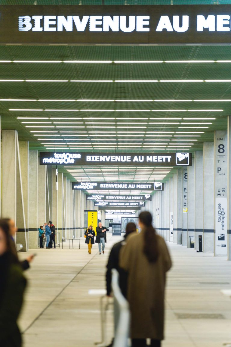 Un long couloir avec des panneaux lumineux indiquant "BIENVENUE AU MEETT". Plusieurs personnes marchent ou se tiennent debout, certaines avec des bagages. L'espace est moderne, avec des piliers en béton, un haut plafond et des panneaux indiquant les halls et les parkings sur le mur de droite.