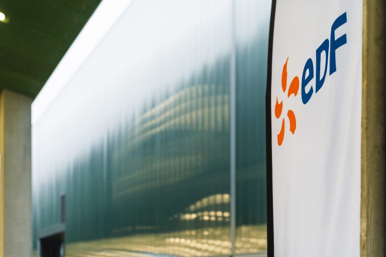 Un gros plan d'une bannière blanche avec les lettres bleues "eDF" et un logo orange en forme de soleil est affiché sur la droite. À l'arrière-plan, un bâtiment moderne en verre teinté vert avec des lignes verticales et des piliers sous un toit vert.