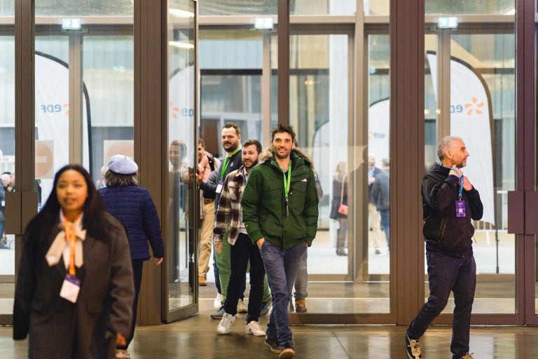 Un groupe de personnes, dont certaines portent des badges et des cordons, franchit de grandes portes vitrées lors d'un événement en salle. L'atmosphère est lumineuse et accueillante, et plusieurs personnes sourient. L'arrière-plan montre d'autres participants et des bannières à l'extérieur.