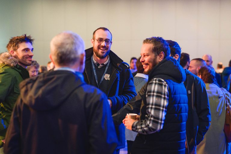 Un groupe de personnes discute lors d'un événement en salle. Deux hommes au premier plan sourient, l'un d'eux tenant une tasse. D'autres sont détournés ou flous à l'arrière-plan, portant des vestes décontractées. L'atmosphère semble amicale et sociale, avec un éclairage chaleureux.