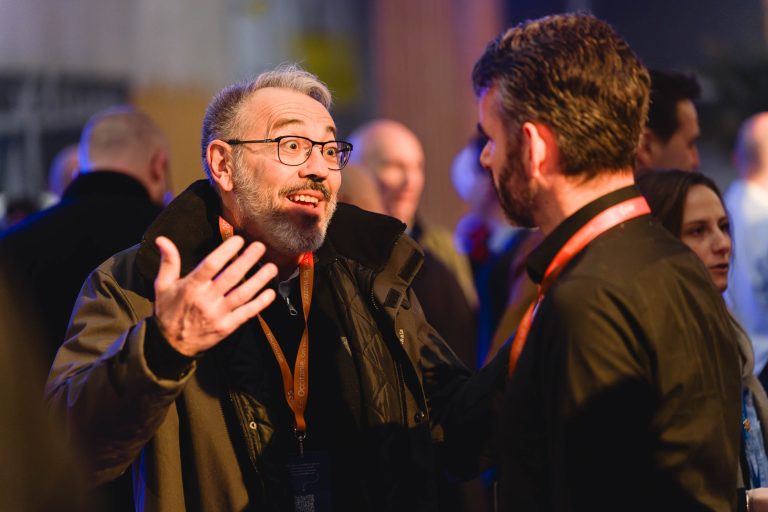 Un homme avec des lunettes et une barbe grise fait des gestes animés avec les mains levées tout en parlant à un autre homme lors d'un événement intérieur bondé. Tous deux portent des cordons et plusieurs personnes sont visibles dans l'arrière-plan à la lumière tamisée. L'éclairage est chaud et ambiant.