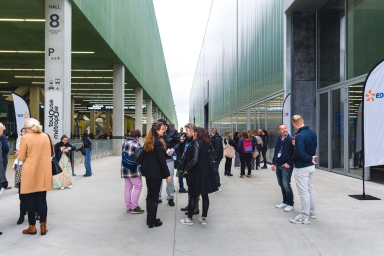 Un groupe de personnes discute à l'extérieur d'un grand bâtiment moderne aux murs de verre et à l'auvent vert. Certains portent des badges et des manteaux. Des drapeaux et des panneaux de signalisation sont visibles, et d'autres personnes marchent à l'arrière-plan. Il semble qu'il s'agisse d'une conférence ou d'un événement.