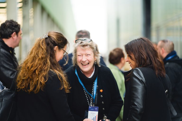 Un groupe de personnes se tient à l'extérieur, discutant et souriant. L'accent est mis sur une femme âgée au centre, avec des cheveux gris et une lanière, qui rit chaleureusement avec deux femmes qui lui font face. L'arrière-plan est flou, suggérant un événement ou un rassemblement en plein air.