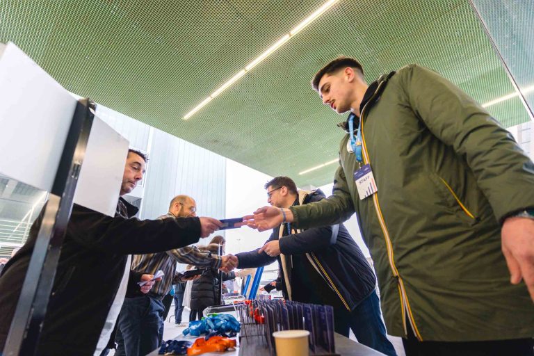 Plusieurs personnes se tiennent à une table d'inscription à l'intérieur, distribuant des badges de conférence et des stylos. Un homme vêtu d'un manteau vert tend un badge à un membre du personnel. La table est équipée de gobelets, de stylos et de cordons bleus. Des plafonniers modernes sont visibles au-dessus de la table.