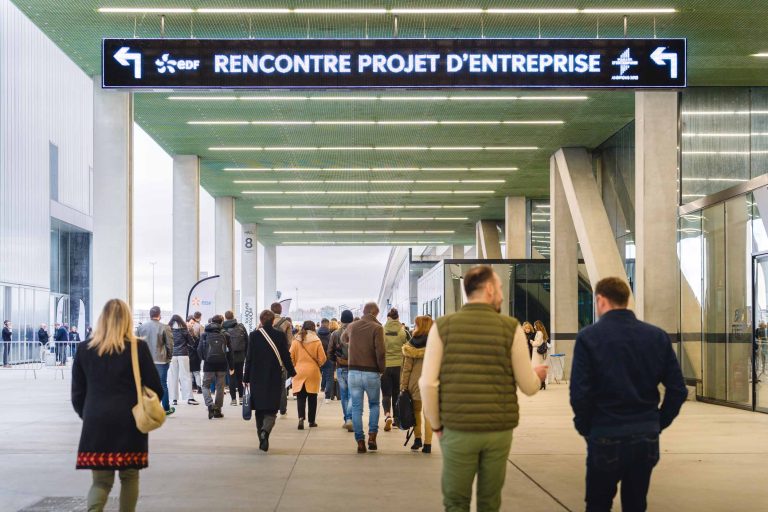 Un groupe de personnes, vêtues de tenues décontractées et d'affaires, se dirige vers l'entrée d'un grand bâtiment sous un plafond vert. Au-dessus d'eux, un panneau numérique en français indique "Rencontre Projet D'Entreprise" avec des flèches directionnelles.