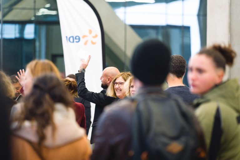 Une femme à lunettes sourit et fait un signe de la main dans un environnement intérieur animé, entourée de personnes engagées dans une conversation. Une bannière blanche avec un texte partiel bleu et orange est visible en arrière-plan, et l'atmosphère suggère un événement social ou de réseautage.
