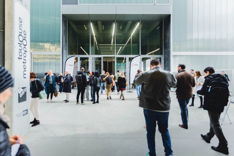 Un groupe de personnes se tient debout et marche devant l'entrée d'un bâtiment moderne aux parois vitrées. Certains parlent, d'autres prennent des photos. Deux drapeaux blancs affichent "Toulouse Métropole". L'atmosphère est animée et décontractée.