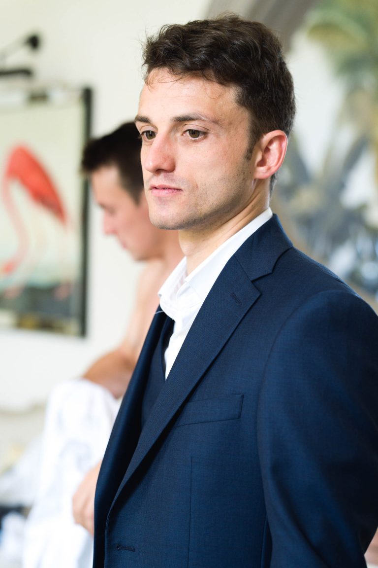 Un homme en costume bleu foncé et chemise blanche se tient à l'intérieur, regardant légèrement vers la droite - peut-être en train de se préparer pour la mariée au Château de Garde à Moulon. Derrière lui, un autre homme s'habille près d'un flamant rose encadré.