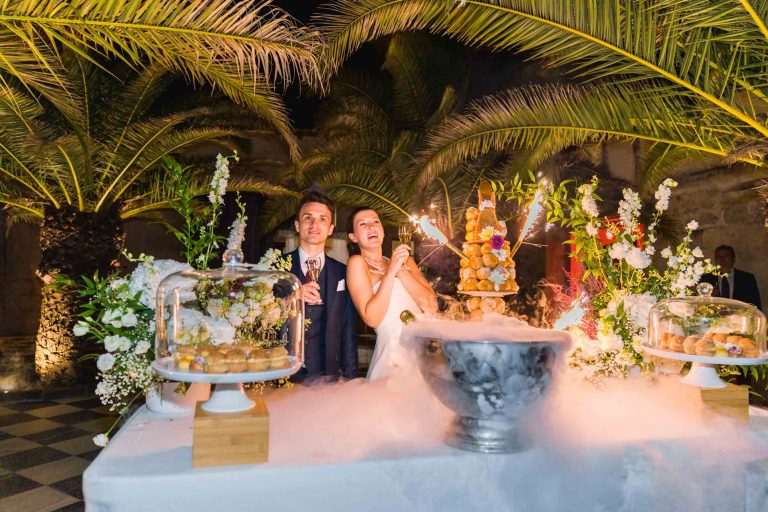La mariée et Antonin sourient derrière une table décorée de fleurs, de pâtisseries et d'un effet spectaculaire de glace carbonique au Château de Garde à Moulon, entourés de palmiers et d'un éclairage chaleureux pour une atmosphère de fête et de célébration.