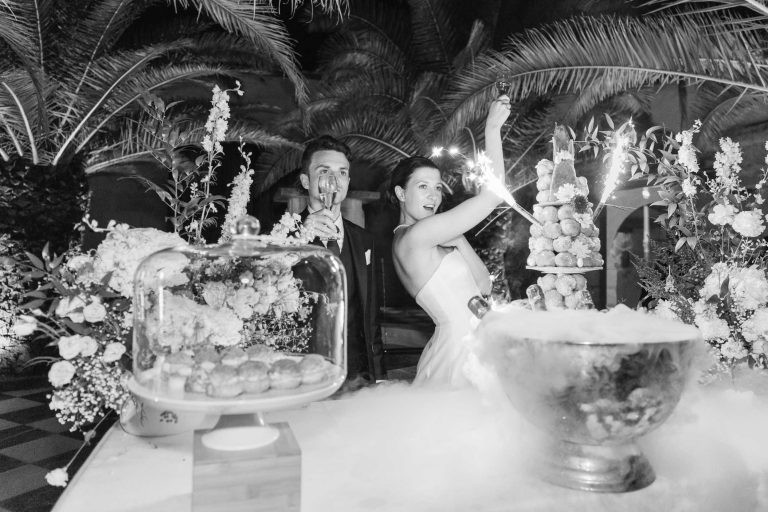 Des mariés, le marié souriant chaleureusement à côté de sa femme, se tiennent derrière une table de fête ornée de fleurs, de pâtisseries sous un dôme de verre et d'un croquembouche. La scène est élégante et joyeuse, parfaite pour une célébration de la Saint-Valentin.