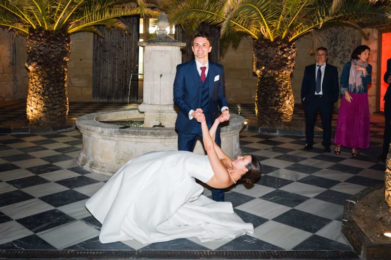 La mariée, vêtue d'une robe blanche, se penche vers l'arrière en tenant la main du marié, vêtu d'un costume bleu marine. Ils dansent sur un sol à carreaux noirs et blancs près de la fontaine en pierre du Château de Garde à Moulon, sous le regard de quatre invités souriants au milieu des palmiers.