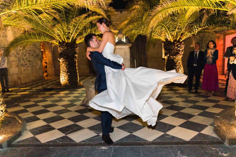 Un marié en costume sombre soulève la mariée dans sa robe blanche fluide alors qu'ils dansent sur un sol à carreaux noirs et blancs en plein air au Château de Garde à Moulon, entourés de palmiers et de murs en pierre chaudement éclairés, sous le regard et le sourire des invités.