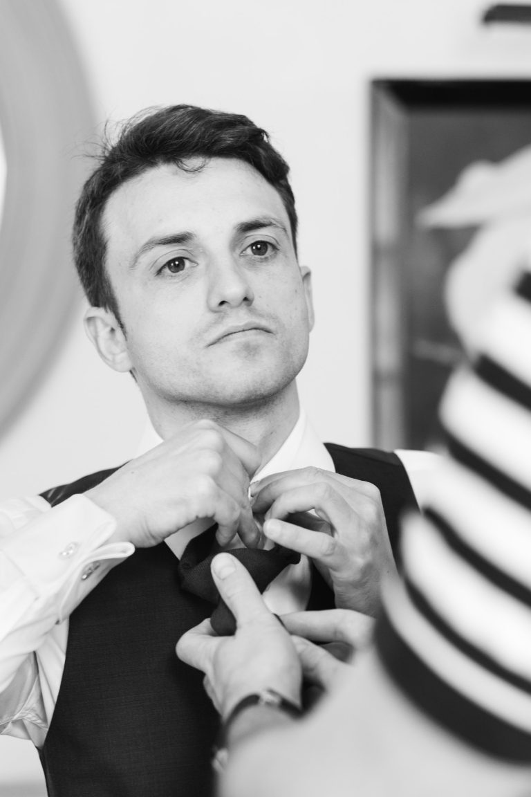 Un jeune homme en gilet et chemise regarde vers le haut tandis que quelqu'un l'aide à ajuster sa cravate - peut-être en train de se préparer pour un événement de la Saint-Valentin à la mairie de Camblanes-et-Meynac. La photo en noir et blanc estompe doucement l'arrière-plan, avec une autre personne en rayures partiellement visible.