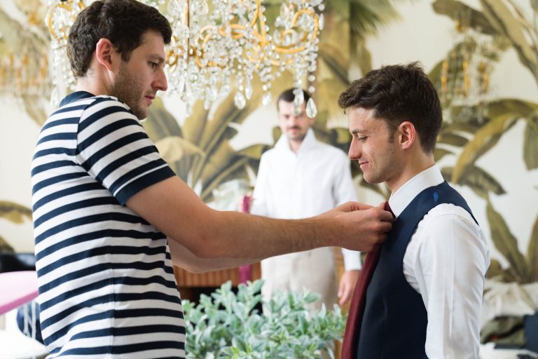 Un homme en chemise rayée aide le marié, vêtu d'une chemise blanche et d'un gilet sombre, à ajuster sa cravate. Un troisième homme se tient dans le flou près d'un papier peint à thème tropical et d'un lustre au Château de Garde à Moulon, créant une atmosphère formelle et élégante.