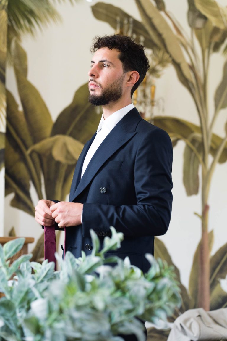 Antonin, un homme aux cheveux bruns courts et bouclés et à la barbe taillée, porte un costume bleu marine et une chemise blanche. Il se tient à l'intérieur de la mairie de Camblanes-et-Meynac, une cravate bordeaux à la main. De grandes plantes à feuilles vertes et un papier peint tropical complètent la scène.