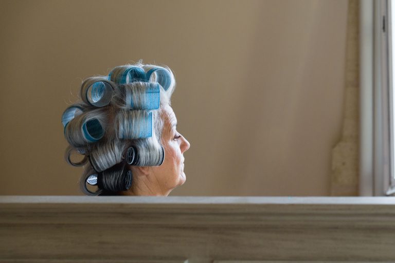 Une femme âgée aux cheveux gris est assise de profil près d'une fenêtre, ses cheveux étant attachés à de gros rouleaux bleus et noirs. Elle semble pensive, placée derrière une surface en bois clair. Le fond beige uni évoque l'élégance tranquille du Château de Garde à Moulon.