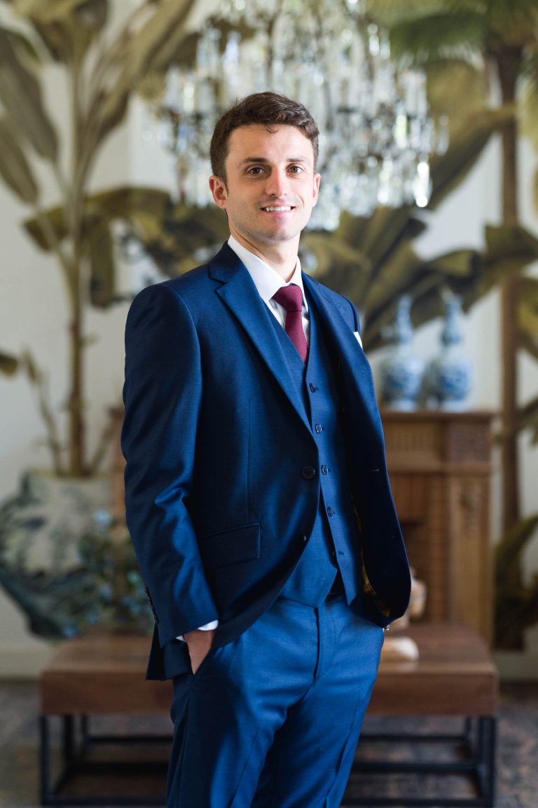 Un jeune homme portant un costume trois pièces bleu marine et une cravate bordeaux se tient à l'intérieur, souriant avec assurance, une main dans la poche. Derrière lui, on aperçoit un papier peint flou à thème tropical, des vases bleus et un lustre en cristal à la mairie de Camblanes-et-Meynac.