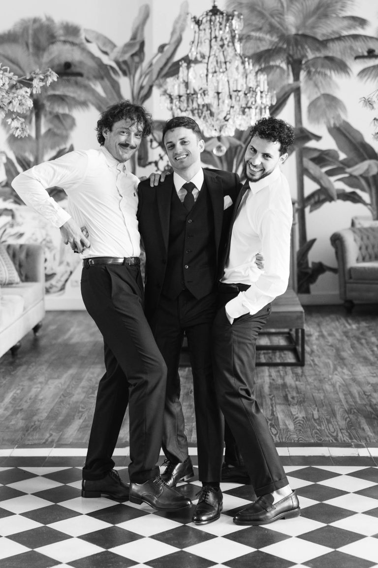 Trois hommes posent ensemble à l'intérieur sur un sol en damier au Château de Garde à Moulon. L'homme du centre porte un costume sombre, flanqué de deux autres en chemise blanche. Tous sourient d'un air enjoué devant un papier peint luxuriant et feuillu, un lustre et des canapés.