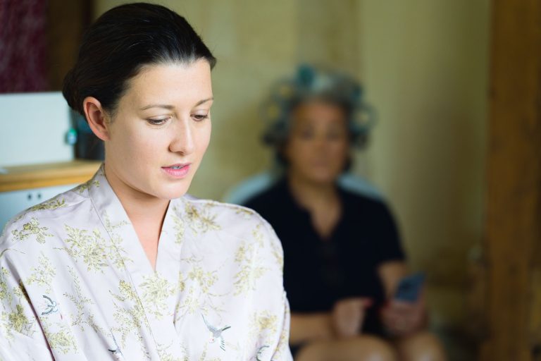 Une femme vêtue d'une robe à fleurs pâles, peut-être la mariée, regarde pensivement vers le bas dans ce décor intérieur calme. À l'arrière-plan flou, une autre femme, coiffée de bigoudis et vêtue de vêtements sombres, est assise et tient un téléphone, son visage n'étant pas net.