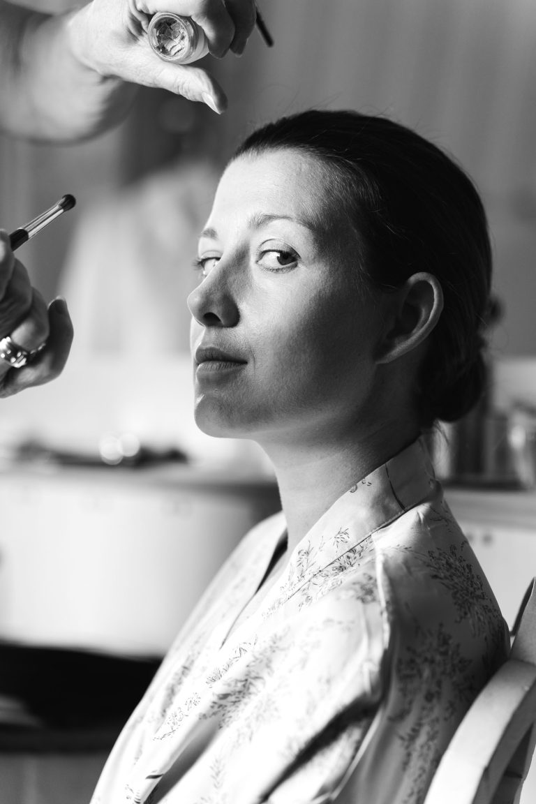 Une femme, la mariée, vêtue d'une robe à fleurs, est assise pendant que quelqu'un lui applique du maquillage sur le visage. Elle regarde calmement et légèrement de côté l'appareil photo. La scène en noir et blanc, doucement éclairée, met en valeur ses traits et l'atmosphère sereine.