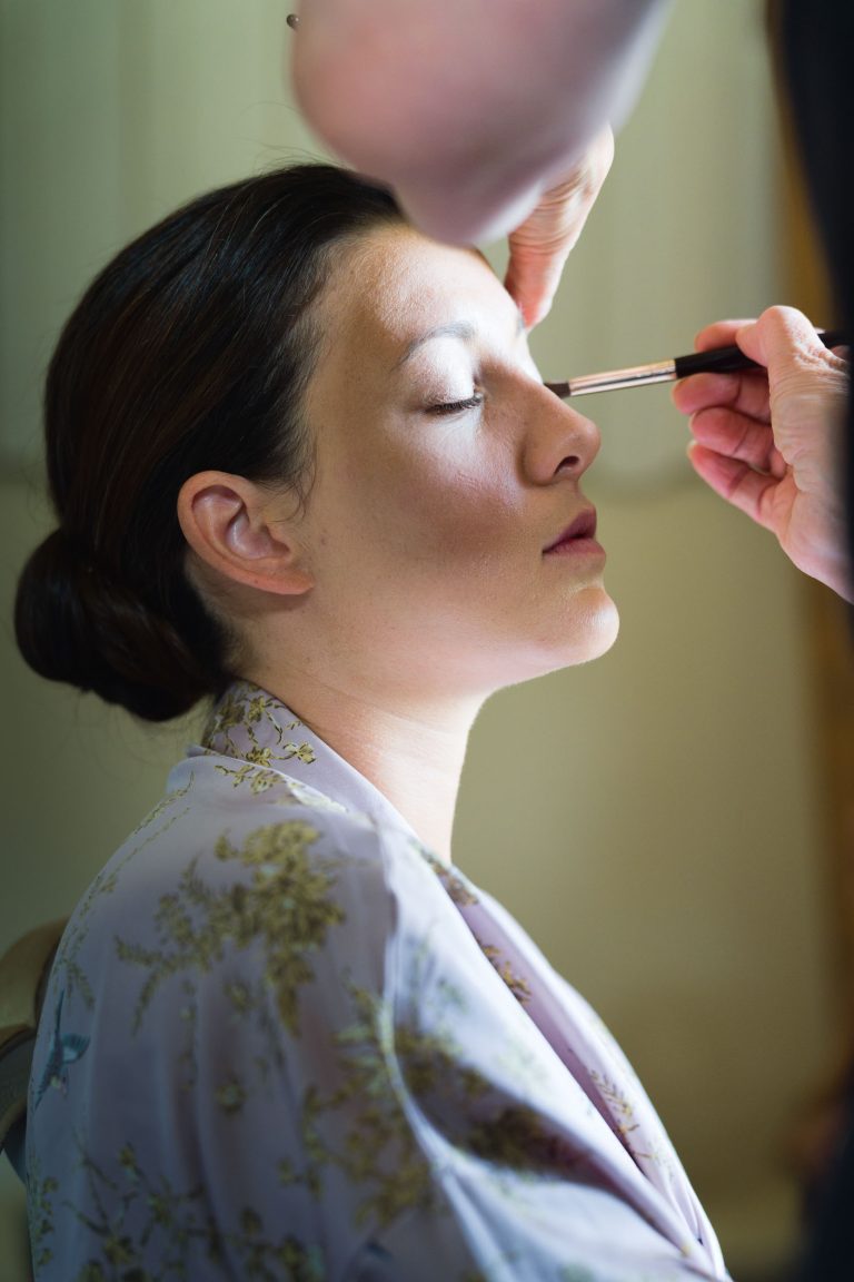 Une femme, la mariée, aux cheveux bruns coiffés en chignon bas et vêtue d'une robe légère et fleurie, est assise, les yeux fermés, tandis qu'une autre applique du maquillage sur sa paupière. La lumière douce et naturelle du Château de Garde à Moulon met en valeur son visage serein et les détails délicats de sa robe.