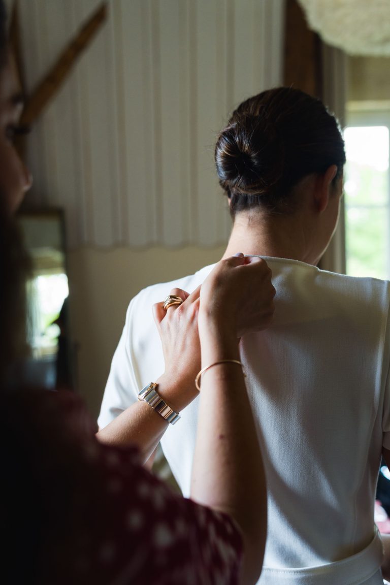 Une femme aux cheveux bruns coiffés en chignon est vue de dos et porte une robe blanche. Une autre personne, partiellement visible, attache le dos de sa robe au niveau de l'encolure. Une lumière douce provenant d'une fenêtre du château de Garde à Moulon remplit l'arrière-plan.