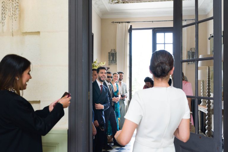 La mariée en robe blanche entre dans une salle lumineuse du Château de Garde à Moulon, face à des invités souriants et bien habillés. Une femme en noir se tient à gauche, en train de prendre une photo. L'atmosphère est joyeuse et la lumière naturelle envahit la scène.