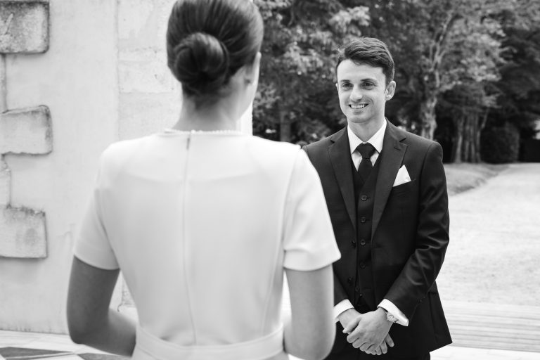 Photo en noir et blanc de Valentin en costume souriant à la mariée, qui se tient debout, les cheveux en chignon. En plein air, près de la mairie de Camblanes-et-Meynac, la scène suggère un événement formel, peut-être un mariage, avec des arbres et un mur de pierre en arrière-plan.