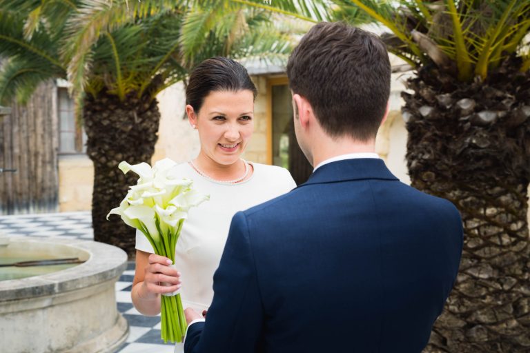 Une mariée en robe blanche tenant des lys calla blancs sourit au marié en costume bleu marine. Ils se tiennent en plein air au Château de Garde à Moulon, près de palmiers et d'une fontaine en pierre, avec un sol en carreaux à carreaux sous eux dans cette scène joyeuse et lumineuse.