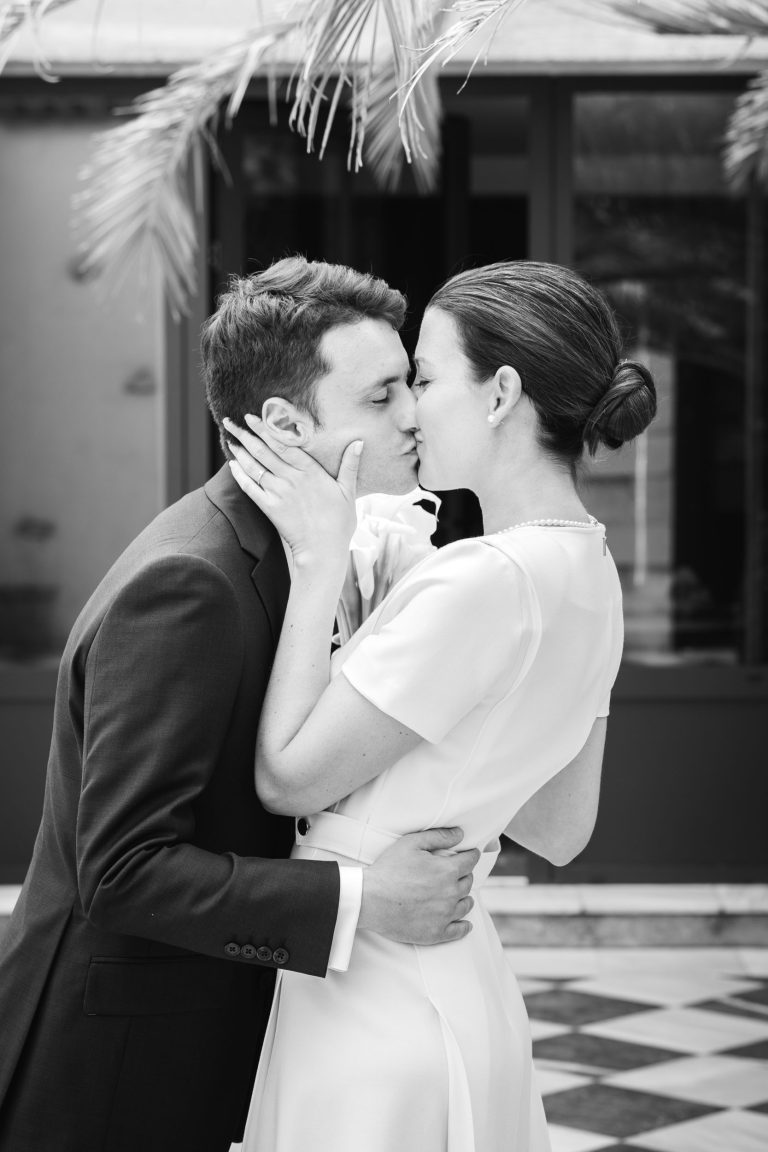 Un couple s'embrasse tendrement en plein air. Le marié, en costume sombre, embrasse la mariée, qui porte une robe claire avec des perles. Sa main se pose sur son visage, la sienne sur sa taille. Des feuilles de palmier et un sol à carreaux apparaissent à l'arrière-plan.