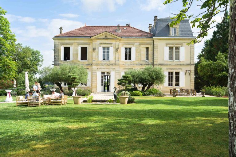 Une grande demeure de couleur claire à deux étages, rappelant le château de Garde à Moulon, se dresse au milieu d'une pelouse verdoyante et de jardins bien entretenus. Plusieurs personnes sont assises et discutent sur des meubles d'extérieur, profitant de la journée ensoleillée encadrée par de grands arbres et arbustes.