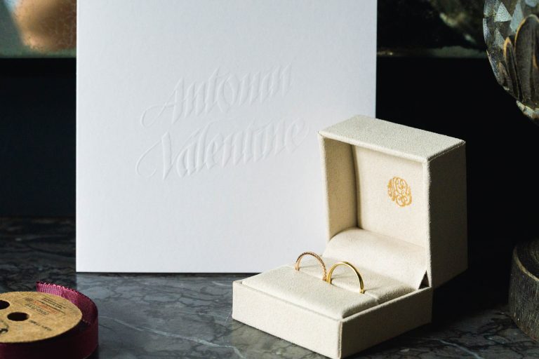 Une boîte à bagues de couleur crème avec deux bagues en or est posée sur une surface en marbre. Derrière, une carte blanche gaufrée indique "Antonin Valentine". Cette scène élégante, à la lumière tamisée, évoque le romantisme d'une Saint-Valentin au Château de Garde à Moulon.