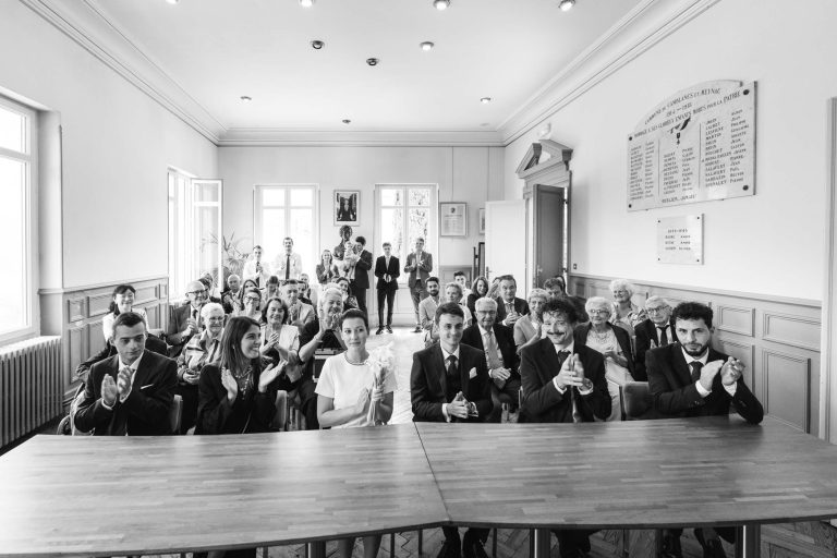 Photo en noir et blanc de personnes habillées de manière formelle, applaudissant et souriant dans une salle lumineuse du Château de Garde à Moulon. Certains se tiennent près de grandes fenêtres. Le cadre suggère que la mariée est célébrée lors de cet élégant mariage ou événement formel.