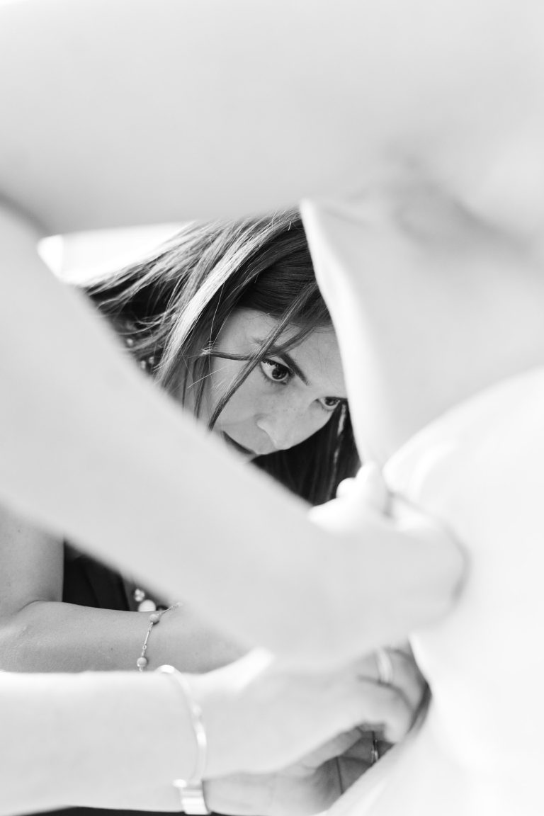 Une femme aux cheveux lisses regarde attentivement tout en aidant à attacher le dos de la robe blanche de la mariée. Son visage est encadré par les bras d'une autre personne dans cette image intime en noir et blanc, peut-être prise avant une cérémonie à la mairie de Camblanes-et-Meynac.