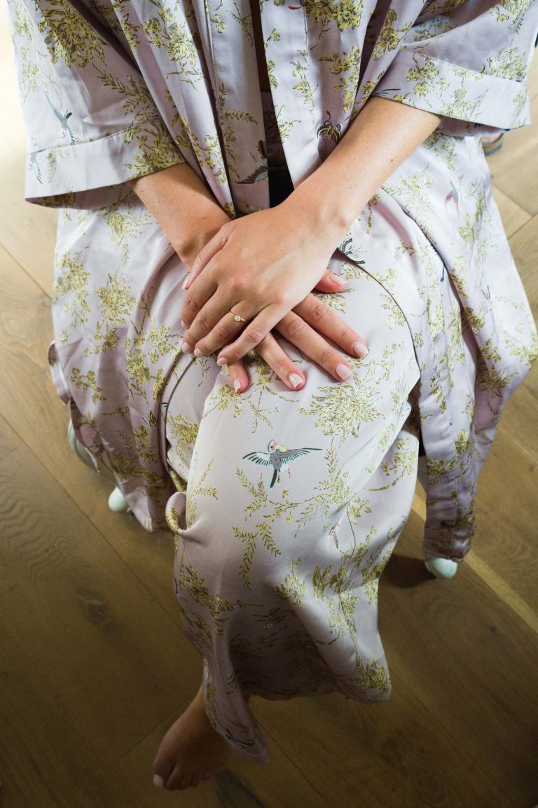 Une personne vêtue d'une robe de chambre à fleurs lavande pâle de Valentine est assise, les mains délicatement croisées sur ses genoux. Ses ongles sont soigneusement manucurés avec un vernis léger, et ses pieds nus reposent sur un sol en bois clair orné de délicats motifs d'oiseaux et de branches.