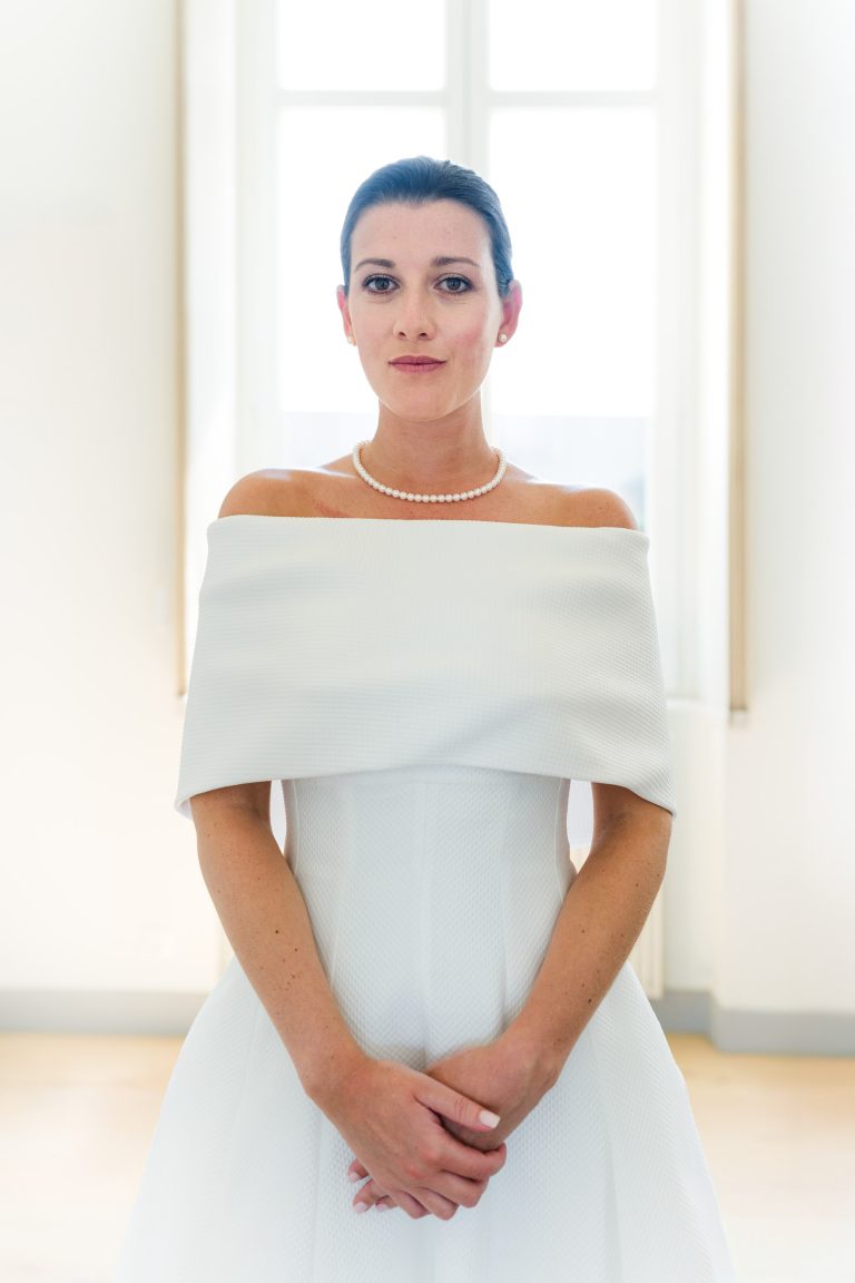 Une femme vêtue d'une robe blanche à épaulettes se tient à l'intérieur du Château de Garde à Moulon, les mains jointes. Elle a les cheveux noirs tirés en arrière, porte un collier de perles et regarde calmement l'appareil photo tandis qu'une lumière douce s'échappe d'une grande fenêtre derrière elle.