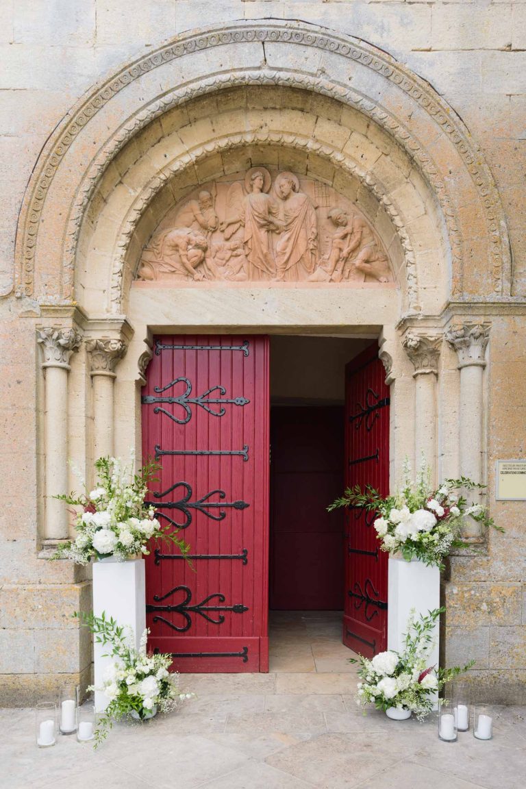 Une entrée d'église en pierre avec des sculptures ornementales au-dessus de doubles portes rouges cintrées à la mairie de Camblanes-et-Meynac. Les portes sont ouvertes, révélant un intérieur sombre orné de compositions florales blanches et de bougies de part et d'autre de la porte.