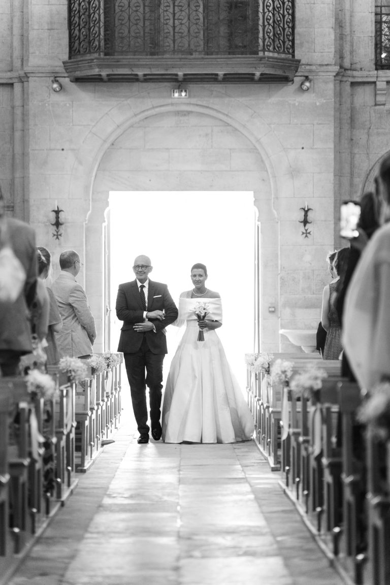 La mariée, vêtue d'une longue robe blanche et tenant un bouquet, descend l'allée avec un homme plus âgé en costume, probablement son père, dans une église. Les invités sont assis de part et d'autre, tandis qu'une lumière vive jaillit de la porte située derrière eux. La scène est en noir et blanc.