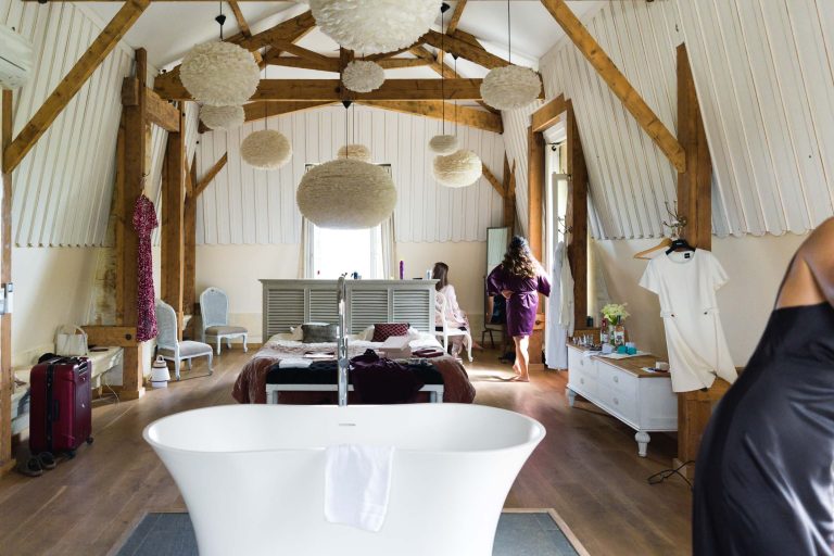 Une chambre spacieuse et rustique avec des poutres en bois, des lampes blanches rondes suspendues et une baignoire indépendante. La mariée, vêtue d'une robe de chambre violette, se tient près de la fenêtre, tandis que les vêtements et les bagages remplissent la pièce aérée - parfaite avant de se rendre à la mairie de Camblanes-et-Meynac.